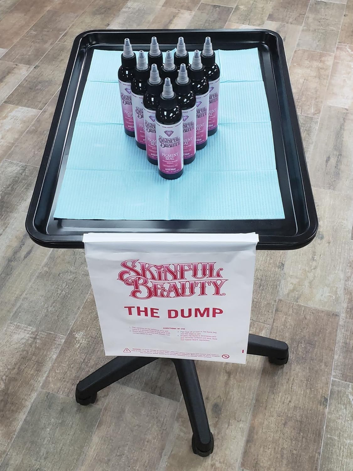 Skinful Beauty “The Dump” disposal bag attached to a workstation tray with multiple Skinful Beauty Pigment Seal bottles on top, showcasing a clean and sanitary PMU setup using Skinful Beauty products.
